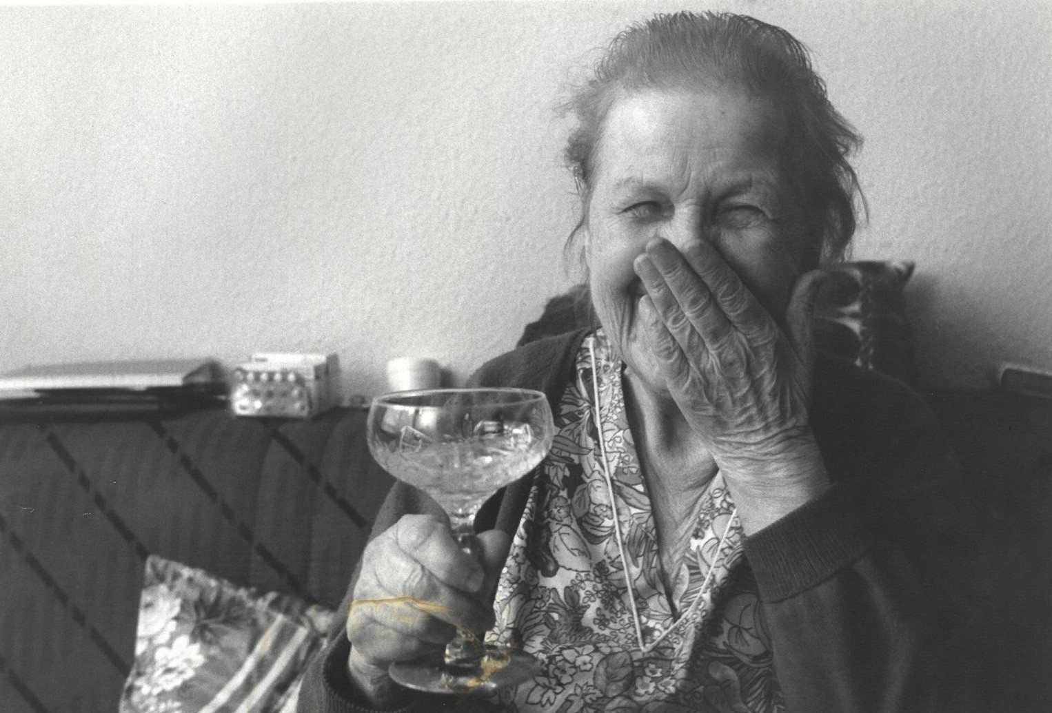 Eine ältere Klientin lacht herzlich hinter vorgehaltener Hand und hält ein Glas Sekt – ein Moment purer Lebensfreude